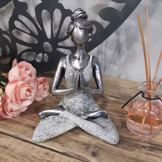 Silver yoga pose figurine on a wooden surface with flowers and a diffuser in the background